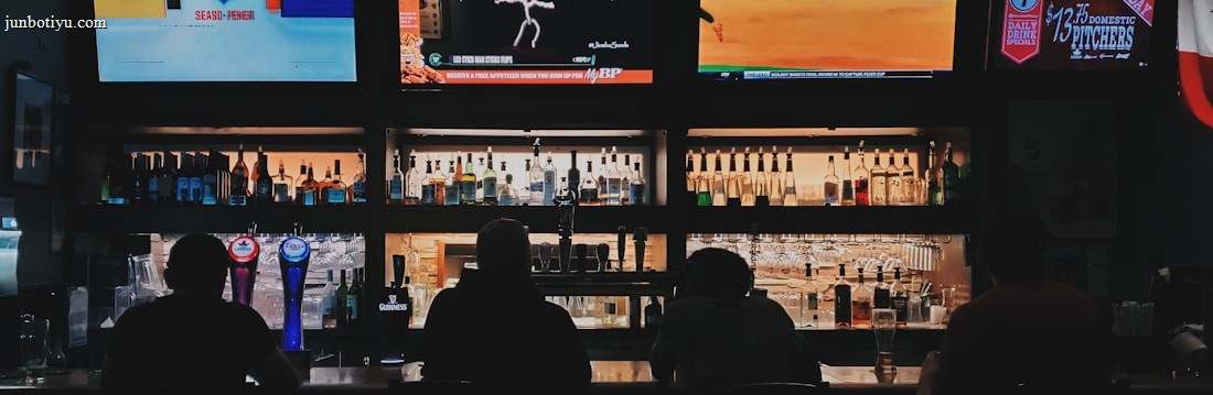 A bar counter with several people sitting in front of it is shown. Above the counter, there are multiple television screens displaying sports broadcasts, including football and baseball. Behind the counter, various bottles of alcohol are neatly arranged.