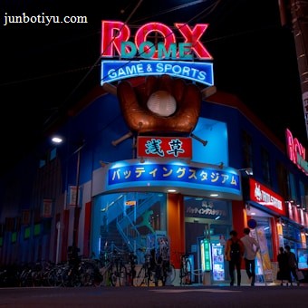 A vibrant, neon-lit facade of a gaming and sports establishment at night, featuring a large glove holding a baseball above the entrance. The building is adorned with colorful signs in both English and Japanese, with various advertisements and an illuminated sign that says 'ROX DOME'. Several people are walking towards the entrance, and bicycles are parked along the side.