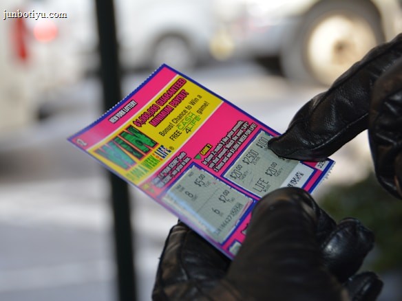 A person wearing black gloves is holding a colorful lottery scratch-off ticket. The ticket includes information about a $1,000,000 guaranteed minimum payout and features multiple games with various prize amounts.